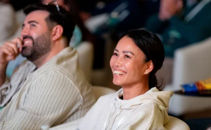 Two people sit indoors, smiling and looking toward the front, appearing engaged in what they are watching or listening to.