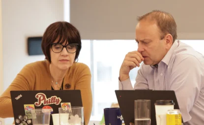 Two people sit at a table using laptops, focused on their screens. Various drinks and items are on the table in front of them.