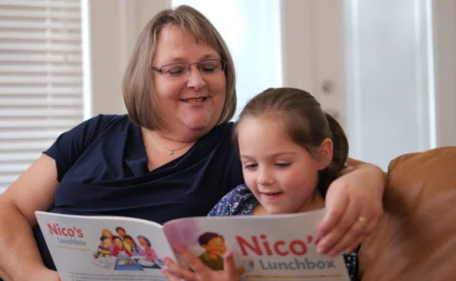 An adult and a young girl sit on a couch reading a children's book titled "Nico's Lunchbox" together, both smiling.