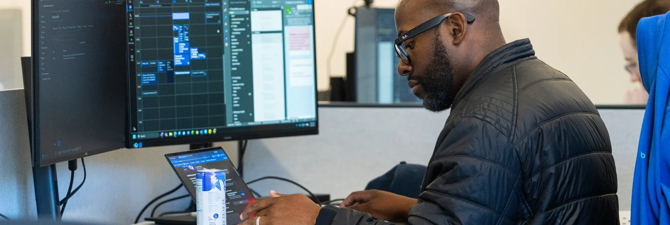 Man wearing glasses and a black jacket works at a desk with a laptop and two large monitors displaying software tools. A beverage can is on the desk.