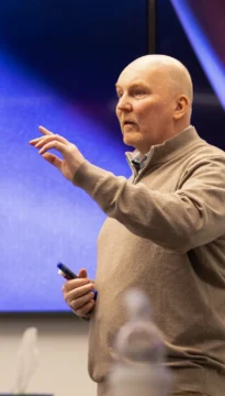 A man in a beige sweater holds a marker and gestures with his hand while speaking in front of a blurred blue background.