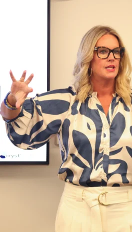 A woman with blonde hair and glasses, wearing a blue and white patterned blouse, gestures with one hand while speaking in front of a screen during a presentation.
