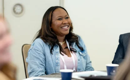 A smiling woman wearing a denim jacket and a conference lanyard sits at a table during a meeting.